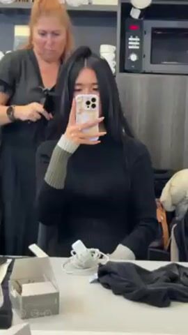 The video shows two women in a hair salon or similar setting. One woman, with auburn hair tied back and wearing a dark dress, is straightening the hair of a younger woman with long, straight black hair. The younger woman is wearing a black turtleneck sweater with gray and green striped cuffs and is holding a cell phone in front of her, seemingly taking a selfie. The phone case has a shimmery, iridescent design. The stylist is using a hair straightener and comb. A makeup table is in front of the younger woman, cluttered with items like a box, a phone charger, and a dark-colored piece of clothing. In the background, there's a microwave, some cups, and a dark-colored wall.
