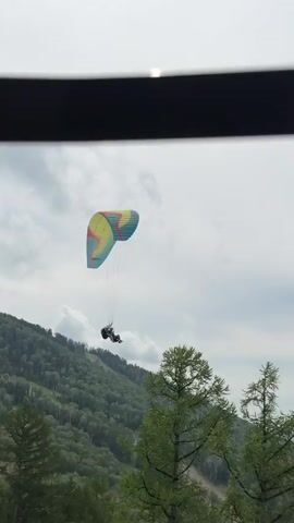 The video captures a paraglider in flight against a cloudy sky. The paraglider is colorful, with sections of yellow, blue, and pink. A person is suspended beneath the paraglider, seated in a harness. The paraglider is positioned above a tree-covered mountainside, with lush green foliage visible on the slopes. The video appears to be filmed from a slightly elevated perspective, possibly from a window or balcony. The overall atmosphere is one of outdoor adventure and exploration.