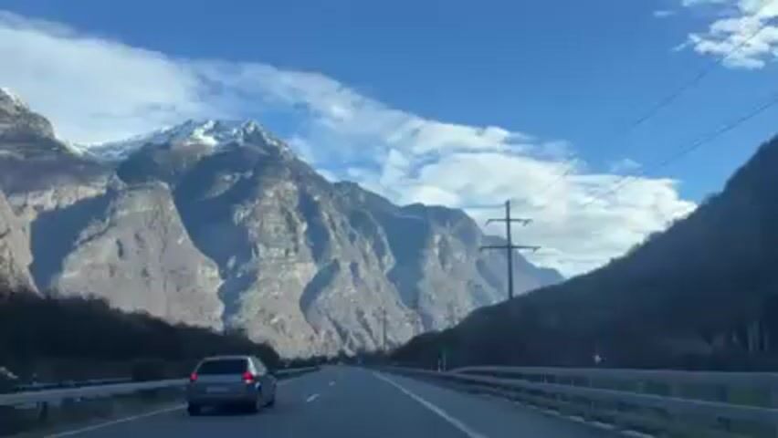 The video shows a view from a moving car driving on a highway with mountains in the background. The sky is blue with some white clouds. A gray car is driving ahead in the same lane. Power lines run parallel to the road. The mountains are tall, rocky, and partly covered with snow at their peaks. The sides of the road are lined with trees and shrubs. The road has clear lane markings.