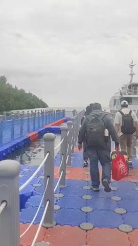 The video shows several people walking down a floating walkway towards a large white boat. The walkway is made of interconnected blue and red modular plastic pieces with a white rope railing on either side. The people are carrying backpacks and luggage. One person in the foreground is wearing a black t-shirt and grey pants, with a black backpack. Another person ahead of them is wearing a white t-shirt and khaki pants, also with a backpack and pulling a red rolling bag. In the background, the large white boat is docked, and there are other smaller boats and a body of water. The sky is overcast.