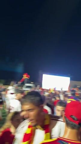 The video shows a crowded outdoor event at night, with a large screen in the background displaying bright images. The crowd consists of many people, mostly young adults, wearing red and yellow clothing, many waving Spanish flags. The atmosphere appears to be festive and energetic, with people cheering and dancing. There are stadium-like structures visible in the background, suggesting a sporting event or concert. The lighting is artificial, with some spotlights illuminating the area. The scene is dynamic and filled with movement.