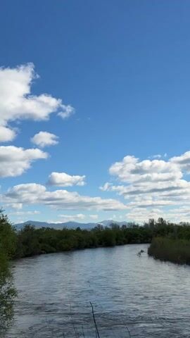 The video shows a scenic view of a river flowing through a lush, green landscape. The river stretches into the distance, flanked by trees and shrubs on both sides. In the background, there is a mountain range with snow-capped peaks. The sky is a vibrant blue, dotted with fluffy white clouds. The overall scene is peaceful and serene. There are no people or animals visible in the shot.