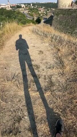 The video opens with a long shadow of a person walking on a dirt path. The shadow is cast by the sun, which is low in the sky, creating a dramatic silhouette. The path is surrounded by dry grass and patches of dirt. In the background, a medieval castle with stone walls and conical towers is visible against a clear blue sky. The camera then pans to show more of the castle, highlighting its imposing architecture and historical significance. The video ends with a wider shot of the castle complex, showcasing its integration with the surrounding landscape. The overall impression is one of a historical exploration, capturing the beauty and grandeur of the ancient fortress under the warm glow of the afternoon sun.