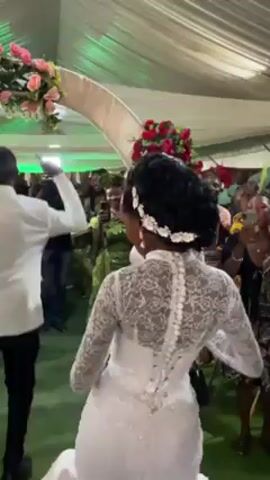 The video captures a lively wedding reception. A bride in a white lace dress with a floral headpiece dances in front of a crowd of guests. They are cheering and recording with their phones. The bride is surrounded by dancing guests. A man in a white suit is seen dancing near her.  The reception is taking place under a white tent with floral decorations. There is an archway decorated with pink and red flowers. The lighting is bright.