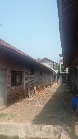 The video shows an outdoor scene with several unfinished or rustic buildings made of brick and wood. The ground is dirt and gravel. There are piles of construction materials, including bricks and wooden planks. A body of water, possibly a pond or canal, is visible in the foreground. The sky is clear and blue, suggesting a sunny day. The buildings appear to be in a rural or semi-rural setting, with some palm trees and other vegetation visible in the background. One building has a corrugated metal roof and exposed wooden rafters. Another building has a wall constructed of large, rough stones. There are some plastic barrels and a ladder leaning against one of the structures. The overall impression is one of a work in progress or a simple, functional construction area.