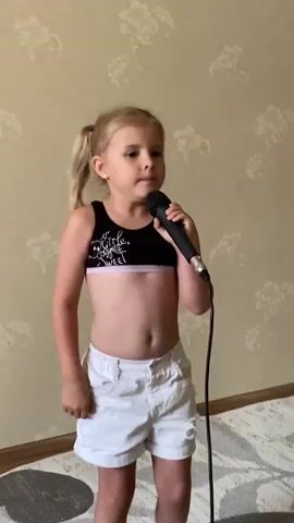 A young blonde girl with pigtails stands in front of a wall with a floral pattern. She is wearing a black crop top with white text and white shorts. She is holding a microphone and appears to be singing or speaking into it. Her mouth is open, and her expression suggests she is engaged in a performance. The background is a wall with a repeating floral wallpaper design. The floor is carpeted with a light-colored, patterned rug. The lighting appears to be artificial, casting a soft illumination on the scene.