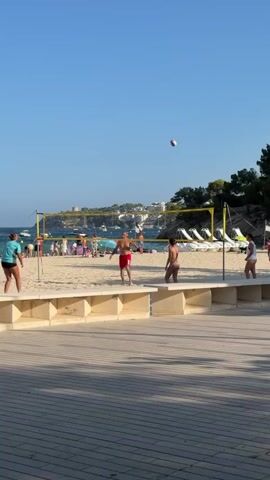 This is a vertical video with a bright blue sky and a beach scene. In the foreground, there is a wooden boardwalk. Behind the boardwalk is a low, beige wall. Beyond the wall is a sandy beach with a volleyball net set up. Several people are visible on the beach, some of whom are playing volleyball. In the background, there is the ocean, with several boats visible, and a coastline with buildings and trees. The video captures a few moments of a volleyball game.