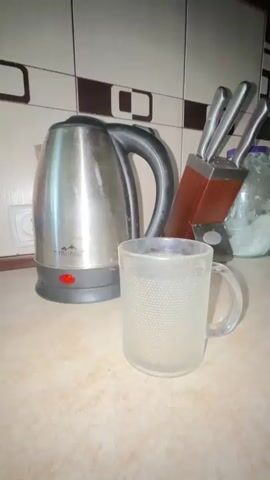 A hand is pouring instant coffee from a red and green packet into a clear glass mug filled with a light brown liquid. A silver electric kettle with a black handle and a red indicator light is visible behind the mug. To the right of the kettle, there is a wooden knife block containing several knives. The scene is set on a speckled countertop in front of a tiled backsplash.