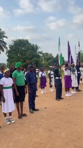 A group of approximately 20 young people, a mix of boys and girls, stand in formation on a dirt field.  Most are wearing similar outfits; some are in blue uniforms with black caps and shoulder stripes, and others are in white and purple skirts with white shirts. A few other people are dressed in green shirts with black skirts or pants. A few hold flags, including the Nigerian flag, at attention. The camera angle captures the group from the side. A young boy in a blue uniform stands facing right, in the foreground. The backdrop consists of a partly cloudy sky, greenery, including trees, and a few buildings in the distance. Someone yells "Sir!" in the background.