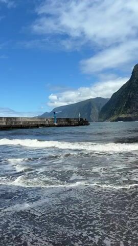 The scene showcases a picturesque coastal landscape, with a black sand beach in the foreground. Gentle waves are lapping at the shore, creating a dynamic yet tranquil feel. A concrete pier juts out into the sea, marked by a crane and a small, striped lighthouse at its end. In the background, majestic mountains rise steeply, covered in lush greenery and fading into the horizon under a bright blue sky dotted with scattered clouds. The overall impression is one of natural beauty and serenity.