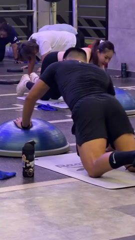 The media file shows a group of people, likely women, exercising in a gym in Nong Pa Khrang, Thailand. The subjects are on their knees on yoga mats, with some using Bosu balls for support. One person in the foreground is on all fours with their hand on a blue Bosu ball. Another person behind them, wearing a black sports bra and black leggings, is also on all fours. Further back, another individual in a white t-shirt and black leggings is performing a similar exercise. The gym appears to be well-lit and equipped with various fitness apparatus in the background. The overall atmosphere suggests a fitness class or group training session. There are no obvious indicators of the time of day or weather. Visible text includes the brand name "Reebok" on a yoga mat and a sticker on a water bottle in the foreground. The individuals appear focused on their exercises.
