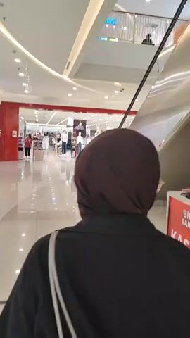 The video shows the back of a person wearing a brown hijab and a black shirt walking through a brightly lit indoor shopping mall. The person is walking towards a large store entrance with red accents and displays of clothing. An escalator is visible to the right. The mall features a modern architectural design with curved white ceilings and recessed lighting. Several other shoppers are visible in the distance, some near the store entrance and others on an upper level.