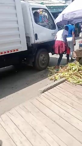 The video shows a bustling outdoor market scene. A white truck is parked on the edge of the pavement, next to a large pile of corn on the cob. Several people are milling around the market stalls, buying and selling goods. One woman in a black and white dress stands at a stall, examining produce. The market stalls are covered by umbrellas, and a sign for "SHREE QUALITY SHOE" is visible in the background.  The pavement is made of wooden planks.