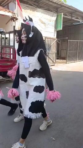 A group of people, mostly young women, are walking in single file down a street. They are all wearing similar costumes resembling cows, with black and white splotches and headpieces with horns. They are also wearing black headscarves and black leggings, with some wearing pink fluffy accessories. The street appears to be in a residential area with houses and buildings on either side. There is a motorcycle parked on the right side of the street, and a few people can be seen watching from the balconies of the buildings. The weather appears to be sunny. The people are walking away from the camera, and the scene is filmed from a slightly elevated perspective.