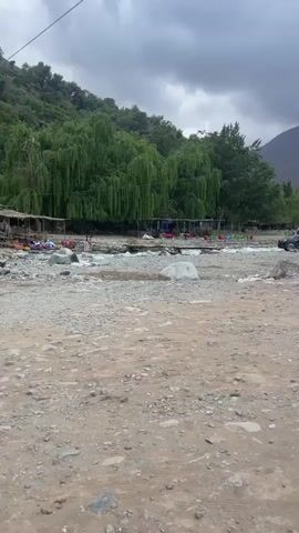 The video presents a series of panning shots showcasing a mountainous landscape in Asgaour, Morocco.

The scene features a wide, rocky riverbed with shallow water, flanked by lush green trees, including prominent willows, on one bank. Several structures, possibly riverside cafes or restaurants with colorful lighting, are visible among the trees. On the near side of the riverbed, the ground is gravelly and dirt.

As the camera pans, a collection of cars, including dark sedans and an SUV, are seen parked across the riverbed and along the banks. In the background, majestic, dark, rocky mountains rise, with their peaks often obscured by heavy, grey clouds, suggesting an overcast day or recent rainfall. A village with multi-story, reddish-brown buildings is built into the lower slopes of the mountains, blending with the rugged terrain. Some buildings feature distinctive architecture.

The sky transitions from dense grey clouds to patches of blue sky with white, fluffy clouds as the camera continues to pan, offering a broader view of the valley. A person is briefly seen walking in the foreground. The overall setting is a natural, rugged environment with elements of human settlement integrated into the landscape. No discernible text is visible.