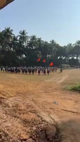 The video shows a group of people gathered on a dirt field, performing what appears to be a choreographed dance or drill. There are at least two red flags planted in the background, slightly off-center from the group. Palm trees surround the field, indicating a tropical or subtropical environment. The sky is clear and blue. Some of the people are wearing white shirts, but their lower garments are dark in color. The perspective is from a slightly elevated position, looking down onto the field. A person can be seen off to the side, facing the group, possibly directing them.