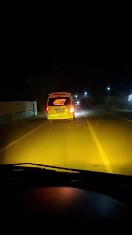 A white van with an orange and red logo on the back is driving down a dark road at night, illuminated by yellow headlights. The van has writing on its back window, and a license plate that is partially visible. The road is paved and has a yellow line down the middle. There are trees on either side of the road, and some lights in the distance. The video is shot from inside another vehicle, looking out through the windshield.