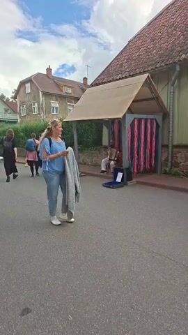 The video shows a street scene that appears to be an outdoor market or festival. In the foreground, a woman with blonde hair is walking and holding a grey blanket over her arm. She is wearing a blue shirt with split sleeves and grey pants, and white sneakers. She is looking to her right. In the background, there are several people walking, as well as some buildings. To the right of the woman, under a covered structure with pink and red curtains, a musician is playing an accordion. Further in the background, other people are seen walking and browsing stalls. The environment appears to be a town or village, with houses and trees visible. The weather seems to be cloudy.