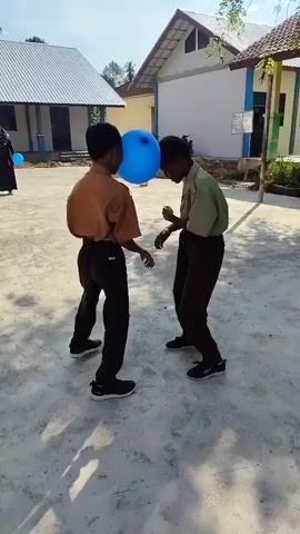 Two young boys, dressed in school uniforms consisting of khaki shirts and dark pants, are playing a game. A large, blue balloon is positioned between their foreheads, and they appear to be pushing against it with their heads. Both boys are facing each other, with their bodies leaning forward in a playful, competitive stance. The ground beneath them is paved with concrete. In the background, two school buildings are visible, one with a white roof and the other with a brown roof and light-colored walls. A person in dark clothing, possibly a hijab, is standing in the distance to the left, also holding a blue balloon. The scene is outdoors, under a bright sky, suggesting a schoolyard or similar open area.
