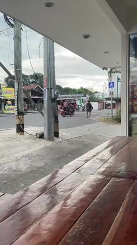 The video shows a street scene from a slightly elevated perspective, possibly from a cafe or restaurant with a wooden table in the foreground. A street with a sidewalk and power lines runs through the middle ground. Several people and vehicles are visible on the street. On the sidewalk, a person is walking away from the camera. Further down the street, a person is walking toward the camera. A red and white tricycle is driving past. A white car is driving away from the camera on the street. Another person is visible near a motorcycle. The sky is overcast.