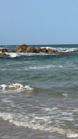 The video presents a still shot of a beach scene, dominated by the interplay of the sea, sand, and rocks. The foreground reveals the shoreline where the light brown sand meets the water's edge, with gentle waves lapping onto the shore. The water transitions from a shallow, foamy white near the beach to a deeper turquoise and blue further out. In the middle distance, a collection of dark brown rocks forms a small, jagged island. Waves are crashing against these rocks, creating white spray and adding dynamic movement to the scene. The sky above is a clear, pale blue, providing a backdrop with no cloud cover. Overall, the scene depicts a tranquil coastal environment under clear skies.