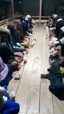 Here is a brief description of the video:

The video shows a group of individuals, mostly women wearing hijabs and niqabs, sitting on a wooden floor indoors. They are arranged in two rows facing each other with small packages and plastic bottles placed in between. The packages are likely food or gifts. The scene is dimly lit and appears to be in a simple, possibly temporary or communal setting. There is chatter and activity among the individuals, suggesting a gathering or event. The surrounding area seems to be an open space with wooden supports and beams.