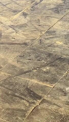 The video shows a close-up of square stone tiles laid on the ground. The tiles are various shades of gray and brown, with some speckled patterns. The sun casts a shadow across the tiles. There are no visible people or other objects in the scene.