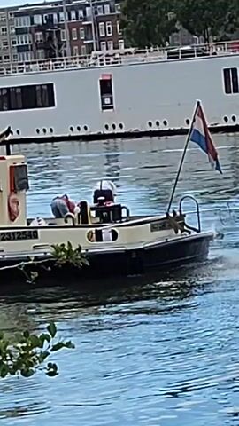 A small tugboat, flying a Dutch flag, navigates on a body of water. A person is visible on the deck of the tugboat. In the background, a large white ship is docked. Buildings and trees can be seen beyond the docked ship. The water is calm and reflects the sky and the surrounding environment. The scene appears to be taking place during daylight hours. The tugboat has the registration number "02312540" visible on its side.