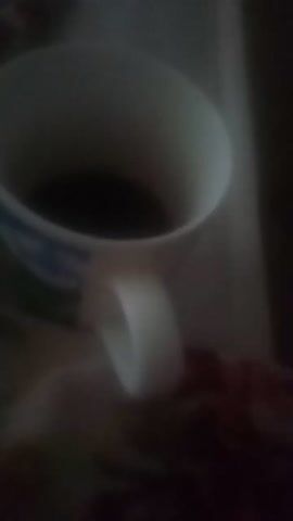 The video begins with a close-up, blurry shot of a white mug with a handle, tilted towards the viewer. The inside of the mug appears dark, possibly containing a beverage. As the video progresses, the mug rotates, revealing a colorful design on its exterior. The design includes a cartoonish orange character with a smiling face, set against a backdrop of a blue sky with white clouds and green hills. The mug also has a blue pattern around its rim. The lighting is dim, and the overall image is out of focus, suggesting the video was filmed in low light conditions or with a low-quality camera. The background is dark and indistinct, making it difficult to discern any specific environmental details. The footage appears to be very short, lasting only a few seconds, and focuses solely on the mug. There are no discernible actions or interactions taking place beyond the rotation of the mug.