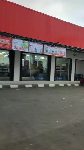 The video begins by showing the exterior of a bright red building with a metal overhang. A sign with colorful images is attached to the overhang. Below the overhang, there are large glass windows displaying rows of blue water bottles. To the right, there is a white cart with a colorful umbrella and a person sitting nearby. The scene then shifts to a wider view of the street, showing more buildings and a road. Another building with a sign that reads "GG Elektronik" is visible, along with several parked motorcycles. The video continues to pan across the street, showing more shops, people, and vehicles, including a white van and a person on a motorcycle wearing a helmet and carrying a box. The environment appears to be an urban street with shops and commercial establishments.