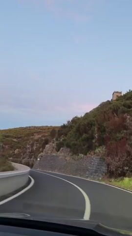 A vehicle drives along a winding, single-lane road in a rural, mountainous area. The road is paved with black asphalt, marked by a solid white line down the center.  The landscape consists of low-lying, scrubby vegetation and rugged rock formations. A gray stone wall runs along one side of the road, followed by a guardrail marked with alternating yellow and black stripes. The sky is light blue, with faint pink hues near the horizon. The video is shot from the driver's perspective inside the car.