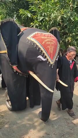 A person wearing traditional clothing walks beside a large, black, elephant-shaped costume. The costume has large ears, tusks, and is decorated with a red and gold cloth on its forehead featuring an elephant emblem and text. The person is holding onto one of the costume's tusks and appears to be guiding it. In the background, a yellow truck is visible with people on top, and trees line the area. The ground appears to be dirt and some paving stones.