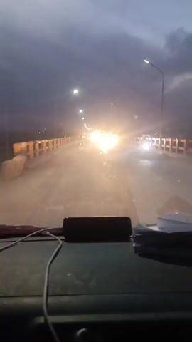 The video, shot from inside a vehicle, shows a nighttime drive across a bridge. The sky is overcast and dark. Streetlights illuminate the bridge. Other vehicles with bright headlights are visible in the distance. A few papers, a black rectangular object, and a white cable rest on the dashboard.