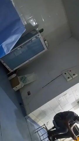 The video begins with a shaky, blurry view of what appears to be a medical facility, possibly an operating room. The camera pans to show a person in dark clothing leaning down, seemingly working near some equipment. The environment is tiled with white and blue colors. The video then shifts to show a patient lying on an operating table, with a medical professional standing nearby, looking at a monitor. Medical equipment, including a ventilator and other devices, are visible. The camera movement is erratic throughout the entire clip.
