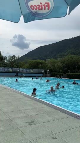 A group of people are swimming in an outdoor swimming pool. The pool is surrounded by a tiled deck and a hedge. In the background, there are trees and buildings, and a mountain covered in trees. The sky is cloudy. The video is shot from a slightly elevated angle, looking down at the pool. The bottom of a blue umbrella with the word "FRISCO" is visible in the top of the frame.