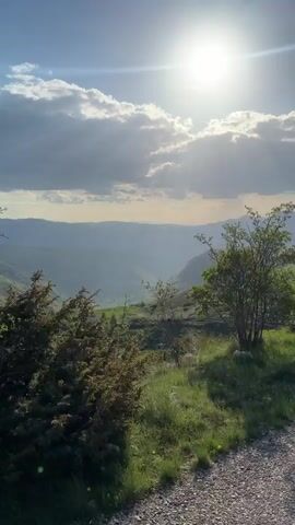 The video shows a mountainous landscape under a partly cloudy, sunny sky. Initially, the sun is high, partially obscured by clouds, and shining down on green hills and valleys. A small tree or bush stands in the foreground. The camera then pans to the left, revealing more of the mountainous terrain, which includes rocky areas and grassy slopes. The sky transitions to a brighter blue with scattered white clouds. A paved path appears in the bottom right corner of the frame, suggesting a road or trail. The video continues to showcase the expansive landscape with its varied terrain and clear sky.