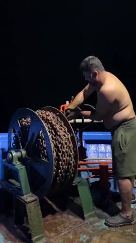 A man, shirtless and wearing shorts, operates a large metal winch laden with thick chain. He is standing on a boat, with dark water and sky visible in the background. He uses his hands to manipulate levers on the winch, suggesting he is either raising or lowering the anchor. The lighting is dim, indicating it is either nighttime or dawn/dusk.