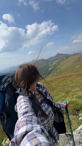 A woman with brown hair in a braid and sunglasses is hiking on a grassy mountain trail. She is wearing a plaid shirt and a backpack. She is holding a hiking pole and smiling at the camera. The sky is blue with some clouds. Other hikers are visible in the distance on the trail. The terrain is hilly and covered in green grass.