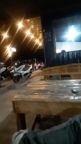 The video shows an outdoor area, probably a restaurant or cafe, at night. The area is illuminated by a string of warm-toned lights hanging overhead. Several motorcycles are parked in a row in front of the structure. The camera is positioned behind a wooden table, partially obscuring the view. The overall lighting is dim, and the scene has a relaxed, informal atmosphere.