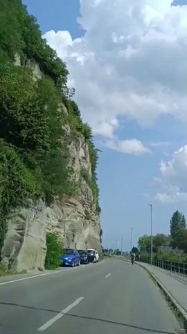 The video shows a road with a rock face on the left and a sidewalk with a railing on the right. There are several parked cars along the rock face, including a blue car and a white car. A person is walking near the white car. Further down the road, there are two cyclists. The sky is blue with white clouds. Trees and vegetation are visible on top of the rock face and on the right side of the road. A body of water is visible in the distance beyond the trees. The video appears to be shot from a moving vehicle.