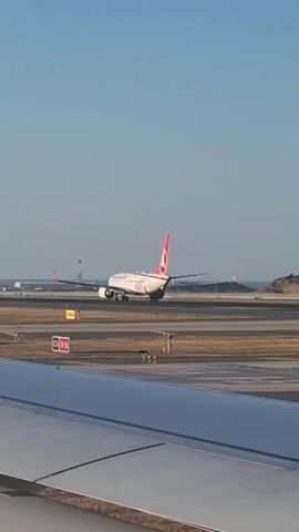 A commercial airplane with the Turkish Airlines logo on its tail is seen on a runway, presumably at an airport. The plane is white with red accents. The sky is clear and blue. In the foreground, a portion of another airplane's wing is visible, suggesting the video was taken from inside a parked or moving aircraft. Other aircraft and airport infrastructure are visible in the background. The lighting indicates it is daytime.