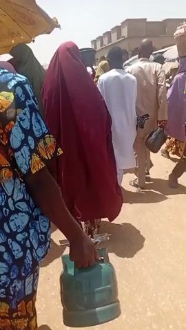 The video depicts a crowded outdoor scene, possibly a market or public gathering. Several people are present, including women wearing traditional Islamic attire. The attire includes hijab-style head coverings in various colors such as burgundy, green, and pink. One individual stands out with a blue shirt patterned with an orange-brown and black design. They also carry a green container. The camera appears to be held by someone within the crowd, capturing the scene at eye level and moving through the people.  Above the crowd, there's a partially visible sunshade with visible writing. Some people in the background are carrying items on their heads. The atmosphere seems busy, with people moving about.