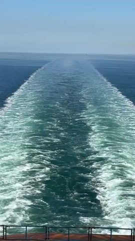 The video shows the view from the back of a ship, looking out over the ocean. The water is a deep blue color, and there are white waves created by the ship's wake. The sky is clear and blue, with a light haze on the horizon. A small sailboat is visible in the distance. The edge of the ship's railing is visible in the bottom of the frame.