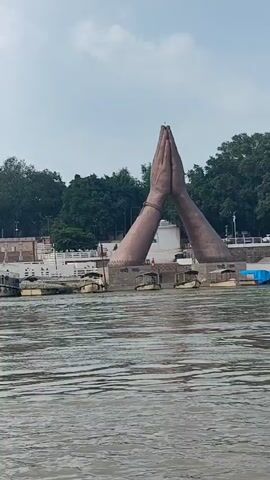 The video shows a wide river with rippling water in the foreground. In the mid-ground, there is a large stone sculpture of two hands in a prayer-like gesture emerging from the water and extending upwards. Behind the sculpture is a white railing along the riverbank, with several boats moored alongside. Further back, there are buildings and lush green trees on a raised embankment. The sky is overcast. There are a few people visible on the riverbank, some walking and some standing near the boats. The overall scene depicts a spiritual or cultural landmark situated by a river.