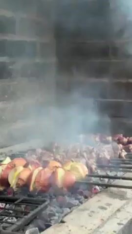 A metal grill sits over a bed of hot coals. Skewers with chunks of meat and onions are grilling, and smoke rises from the food. The grill is set into a brick structure.