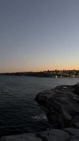 The video shows a view of the ocean and a rocky cliff edge at dusk. The sky is a gradient of deep blue to pale orange near the horizon. The ocean is dark and somewhat choppy. In the distance, a city skyline is visible with lights starting to appear. The rocky cliff in the foreground is rough and textured.