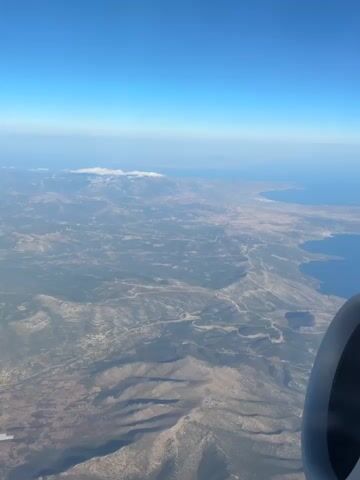 The video shows an aerial view from an airplane window. The landscape below is mountainous and somewhat arid, with patches of vegetation and scattered settlements. A body of water, likely the sea, is visible in the distance, meeting the horizon under a clear blue sky. Part of the airplane's engine cowling is visible in the lower right corner of the frame. The camera appears to be static, capturing a seemingly uneventful flight.