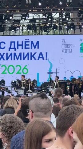The video shows a crowd of people watching a musical performance on an outdoor stage. The banner behind the band reads "Happy Chemist Day! 2026" in Russian. The band members are dressed in black and gold military-style jackets. The crowd is filming and taking pictures of the band with their phones. The stage is well-lit with spotlights.