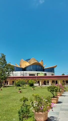 The video shows a building with a unique lotus-shaped roof against a clear blue sky. The building has a yellow facade with a covered walkway. In front of the building is a well-maintained lawn with scattered trees and potted plants. The camera pans slowly across the scene, offering a full view of the exterior architecture and the surrounding landscape. The lighting suggests it is daytime with bright sunlight. There are no people clearly visible or engaged in any significant actions within the frame.