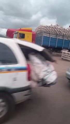 A silver minivan is on the road, moving past a large yellow truck with a red trailer. A blue flatbed truck is behind the yellow truck, carrying a large load of white sacks. A person wearing a headscarf and a red garment is walking away from the blue truck. The road is wet, suggesting it may have rained. Further down the road, houses with corrugated metal roofs are visible, surrounded by trees and vegetation. Another dark-colored car is driving towards the camera. The sky is cloudy.